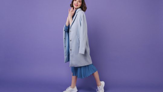 Imagem de uma modelo feminina vestindo um vestido azul com casaco e tênis esportivo, com o fundo roxo