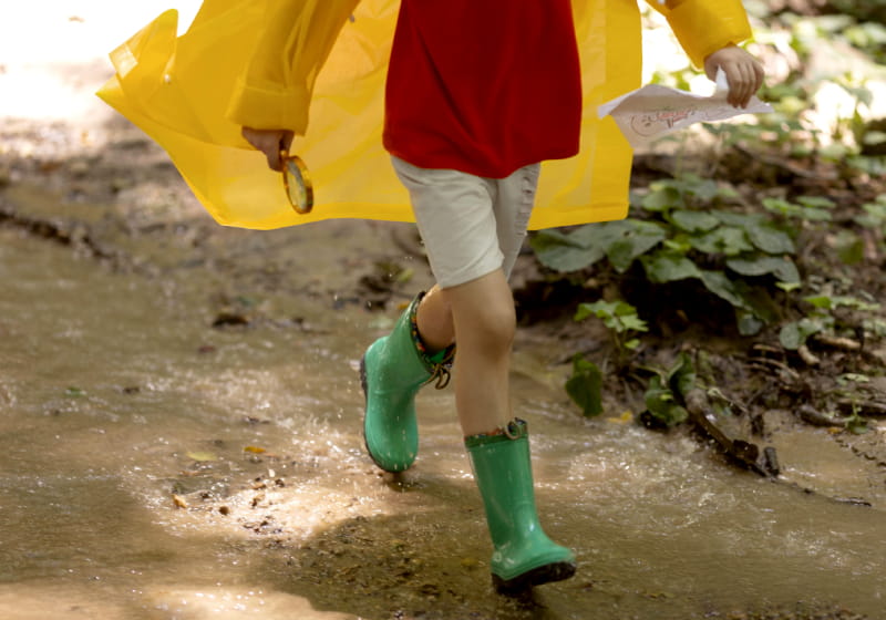 menino andando na poça de água com galocha e capa de chuva com um binoculo em uma das mão e um mapa na outra 