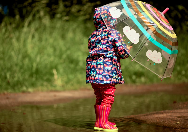 garotinha com guarda chuva na mão com pisando na poça de água com galocha