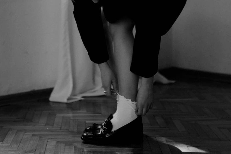 Imagem de uma modelo calçando um par de tamancos pretos com meia cano alto branca, aparecendo da coxa para baixo em uma sala com cortina 