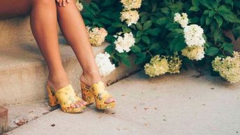 Imagem com uma modelo vestindo um par de tamancos amarelos, aparecendo as pernas, sentada em uma pequena escada ao lado de flores,
