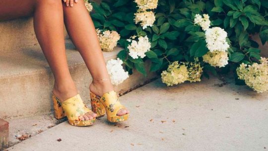 Imagem com uma modelo vestindo um par de tamancos amarelos, aparecendo as pernas, sentada em uma pequena escada ao lado de flores,