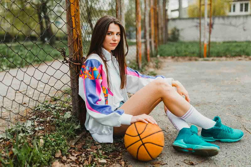 Imagem de uma modelo sentada no chão encostada em uma grade com bola de basquete e tenis azul claro 