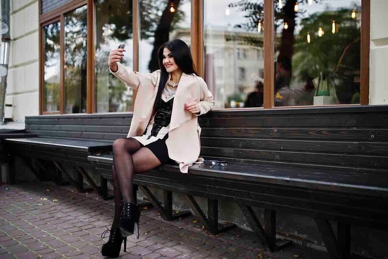 Imagem de uma modelo feminina vestindo uma meia calça preta com vestido preto, salto alto preto e sobretudo, sentada em um banco de madeira em meio urbano, tirando uma selfie com o celular de dia 