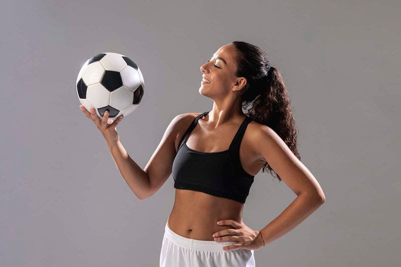 Imagem de uma modelo feminina em pé, vestindo um top preto com shorts branco, com a sua mão esquerda na cintura e a outra segurando uma bola de futebol, com o fundo cinza 