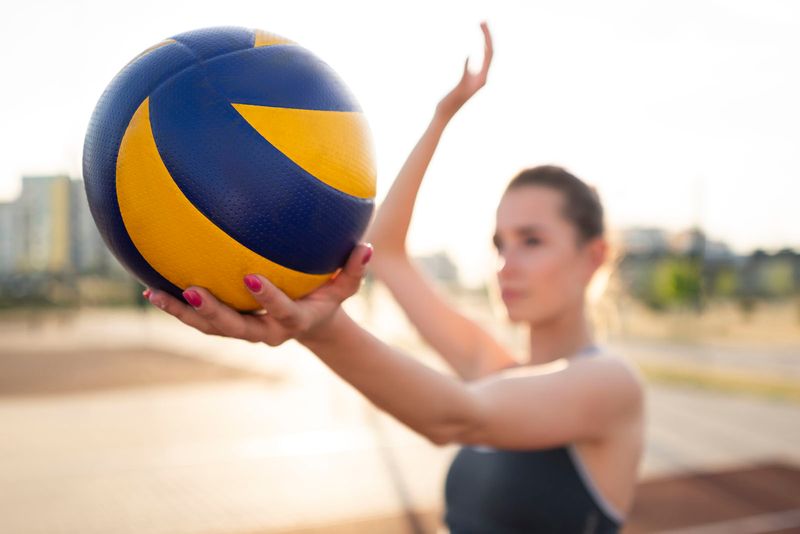 Imagem de uma modelo se preparando para fazer um saque de vôlei, com o fundo desfocado de quadra esportiva, de dia 