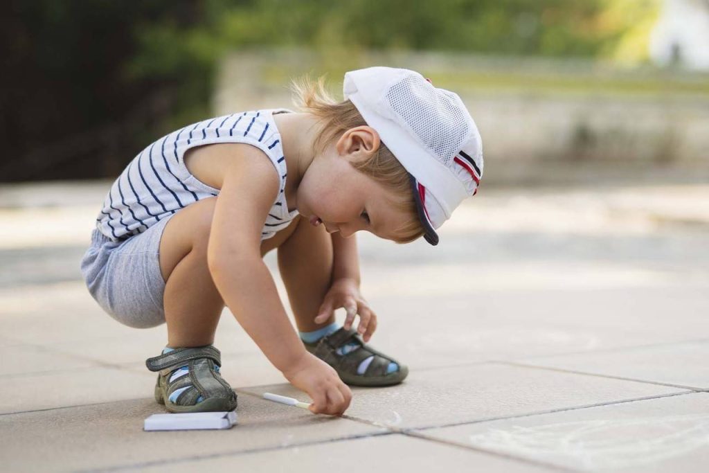 Imagem de um menino, vestindo roupas confortáveis, com um par de tênis, pintando o chão com giz branco. 