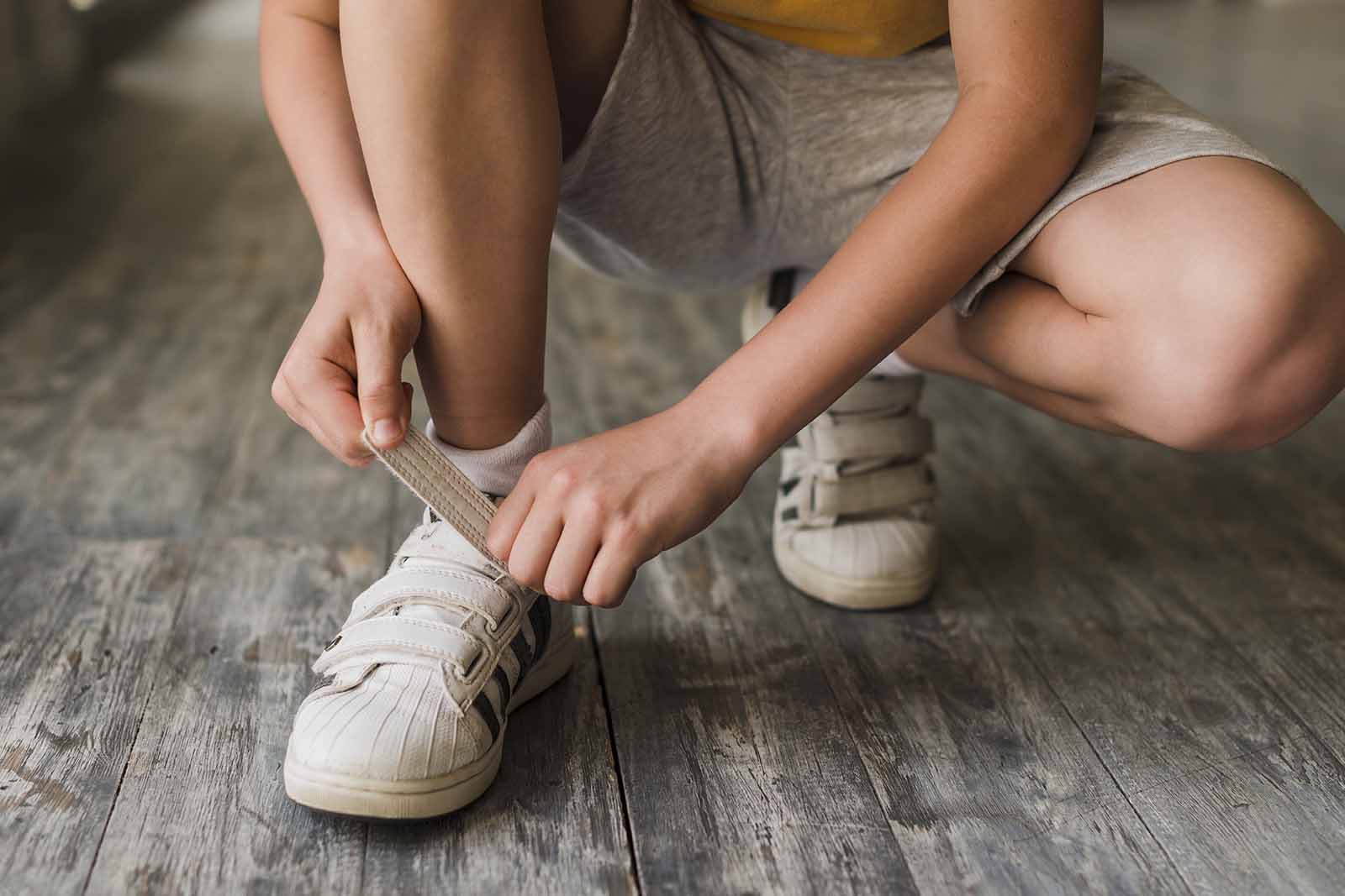 Imagem de um menino, agachado amarrando seu tênis com amarras de velcro, sob um chão de madeira.  