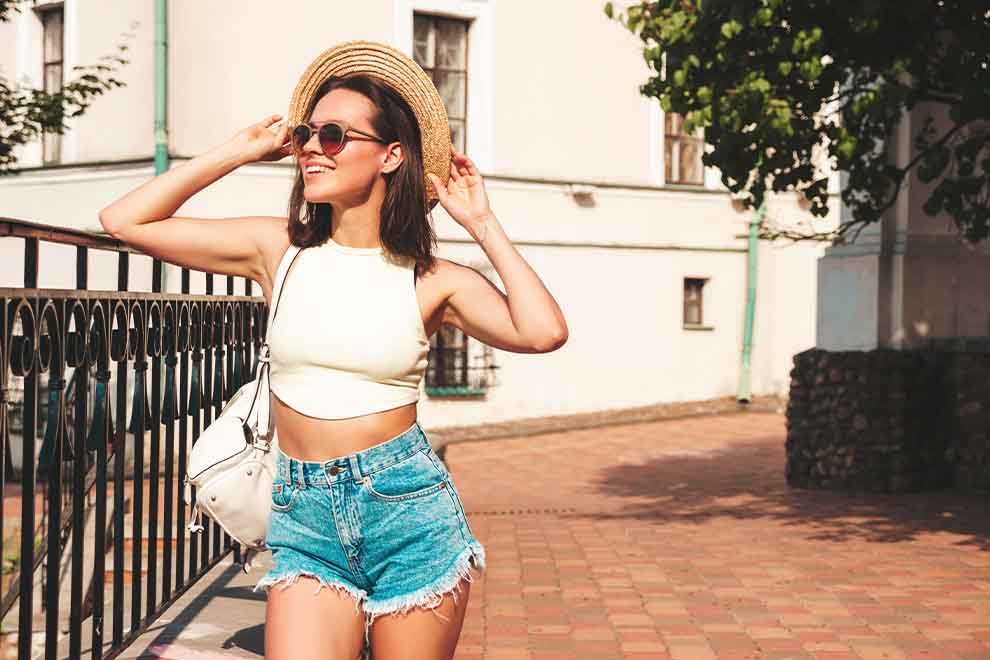 uma mulher usando um cropped branco e shorts jeans com uma bolsa, óculos e chapéu de palha em um dia ensolarado de verão