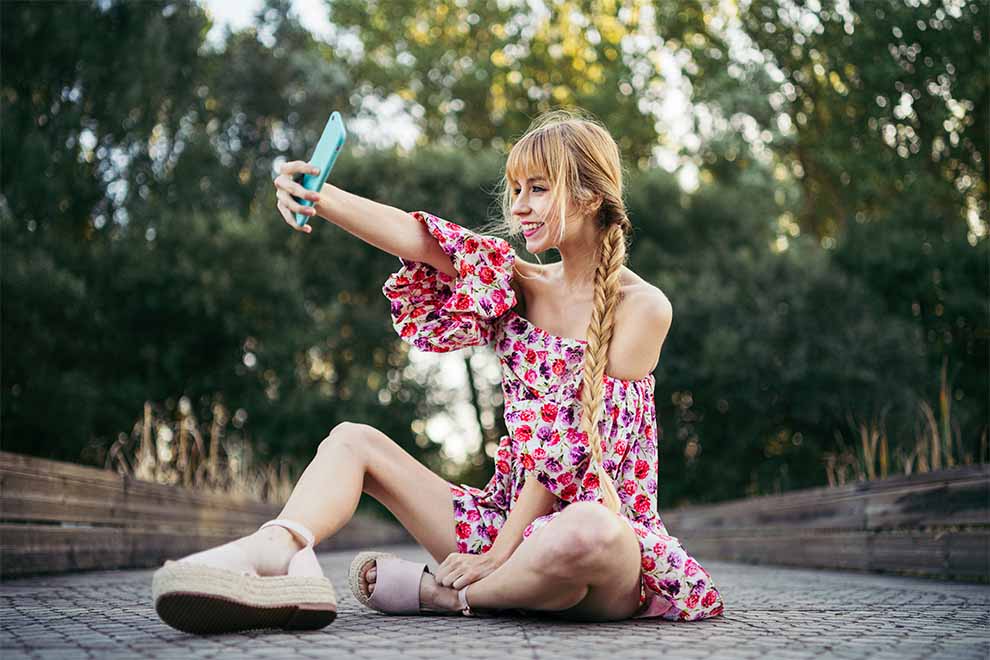 uma jovem usando um vestido solto com um salto anabela sentada no chão tirando uma selfie