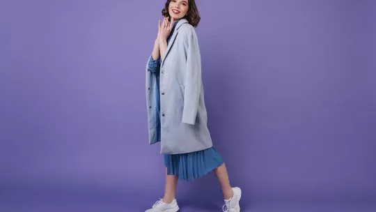 Imagem de uma modelo feminina vestindo um vestido azul com casaco e tênis esportivo, com o fundo roxo