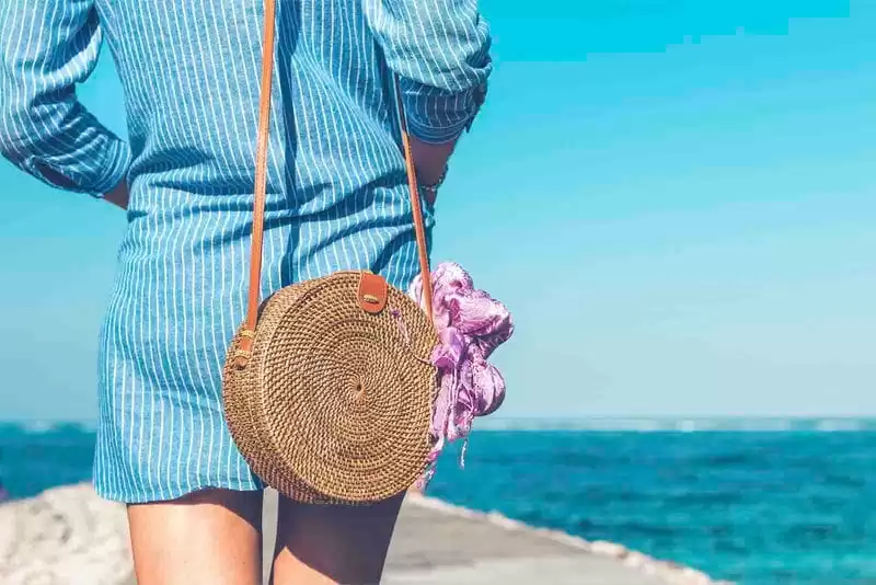 Imagem de uma modelo de costas vestindo uma camisa listrada azul e branca, usando uma bolsa de ombro redonda com alça de couro, em um fundo de mar 
