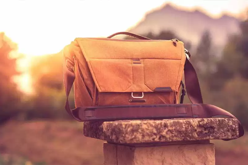 Imagem de uma bolsa transversal marrom  sob um palanque de  pedra com o fundo de arvores desfocadas no por do sol 