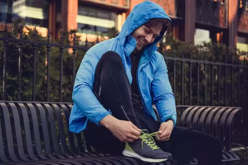 Imagem de um modelo de casaco azul, sentado em um banco de praça amarrando o tenis 