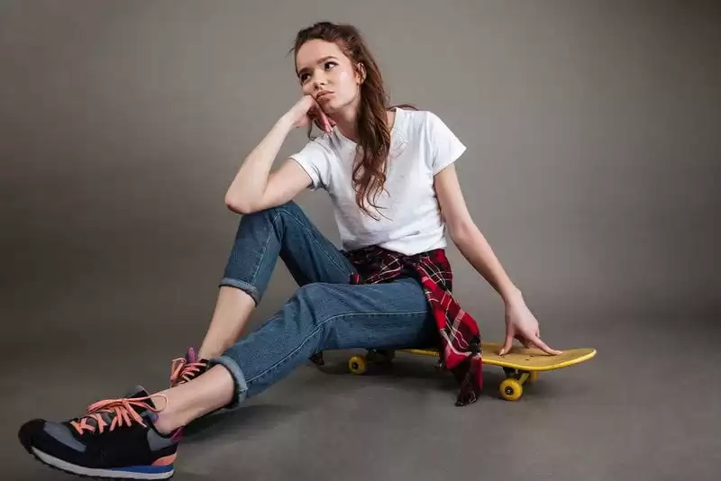 Imagem de uma modelo de calça jeans e camiseta branca, sentada em um skate num fundo cinza escuro 