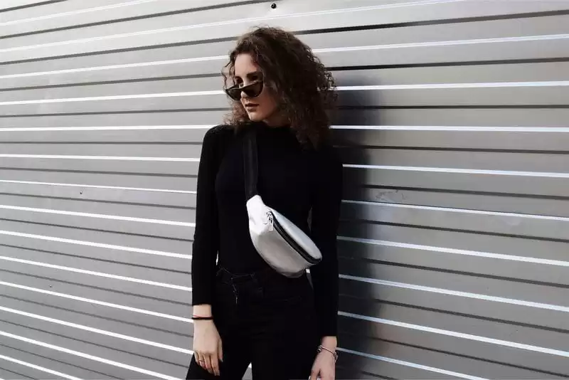 Imagem de uma modelo de frente usando roupas pretas, com uma pochete prata na parte superior do corpo em um fundo de parede metálica cinza 