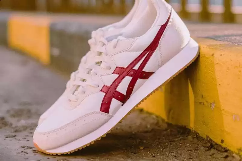 tênis Asics branco com detalhes vermelho inclinado no meio fio de uma rua 